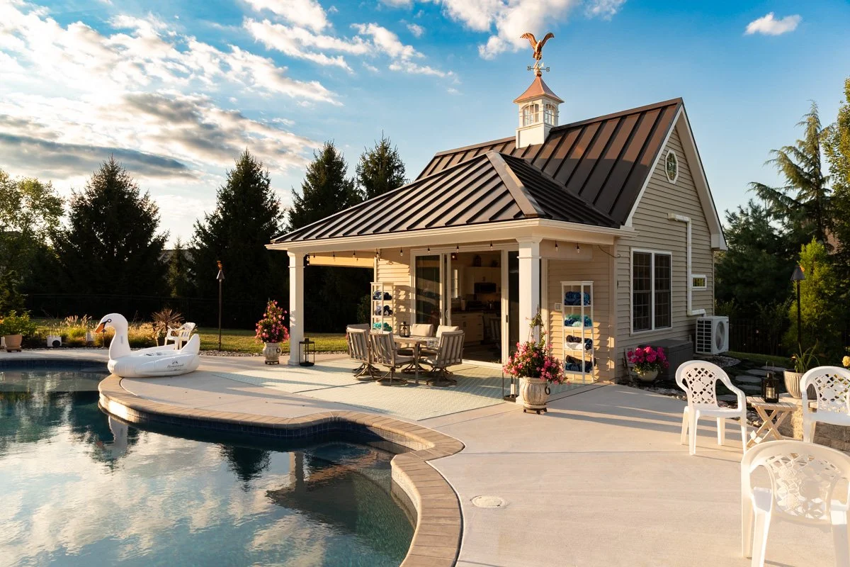 A backyard with a swimming pool, a small house with a metal roof, outdoor furniture, potted flowers, and a floating swan pool float, all under a partly cloudy sky.