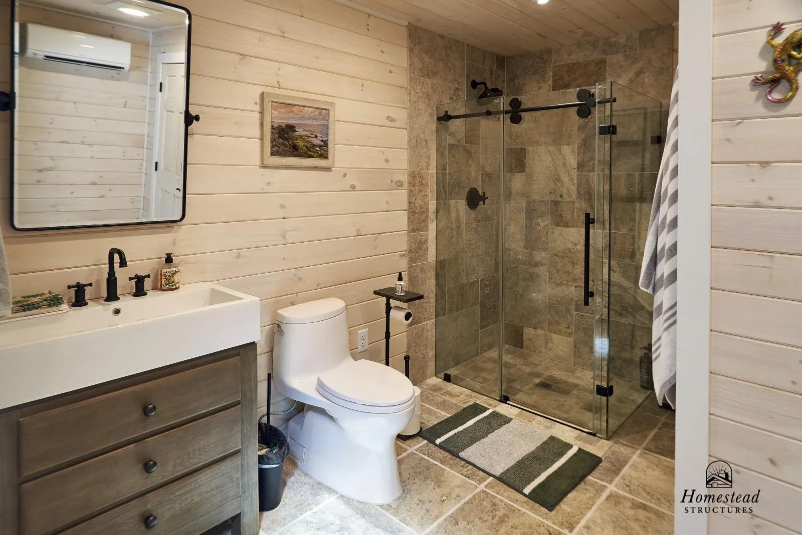 Bathroom with wood-paneled walls, a rectangular mirror, a white sink with black fixtures, a framed landscape painting, a toilet, a small black side table with soap and toilet paper, a glass-enclosed walk-in shower with brown tile, a striped towel hanging on a rack, and a rug on the tiled floor.