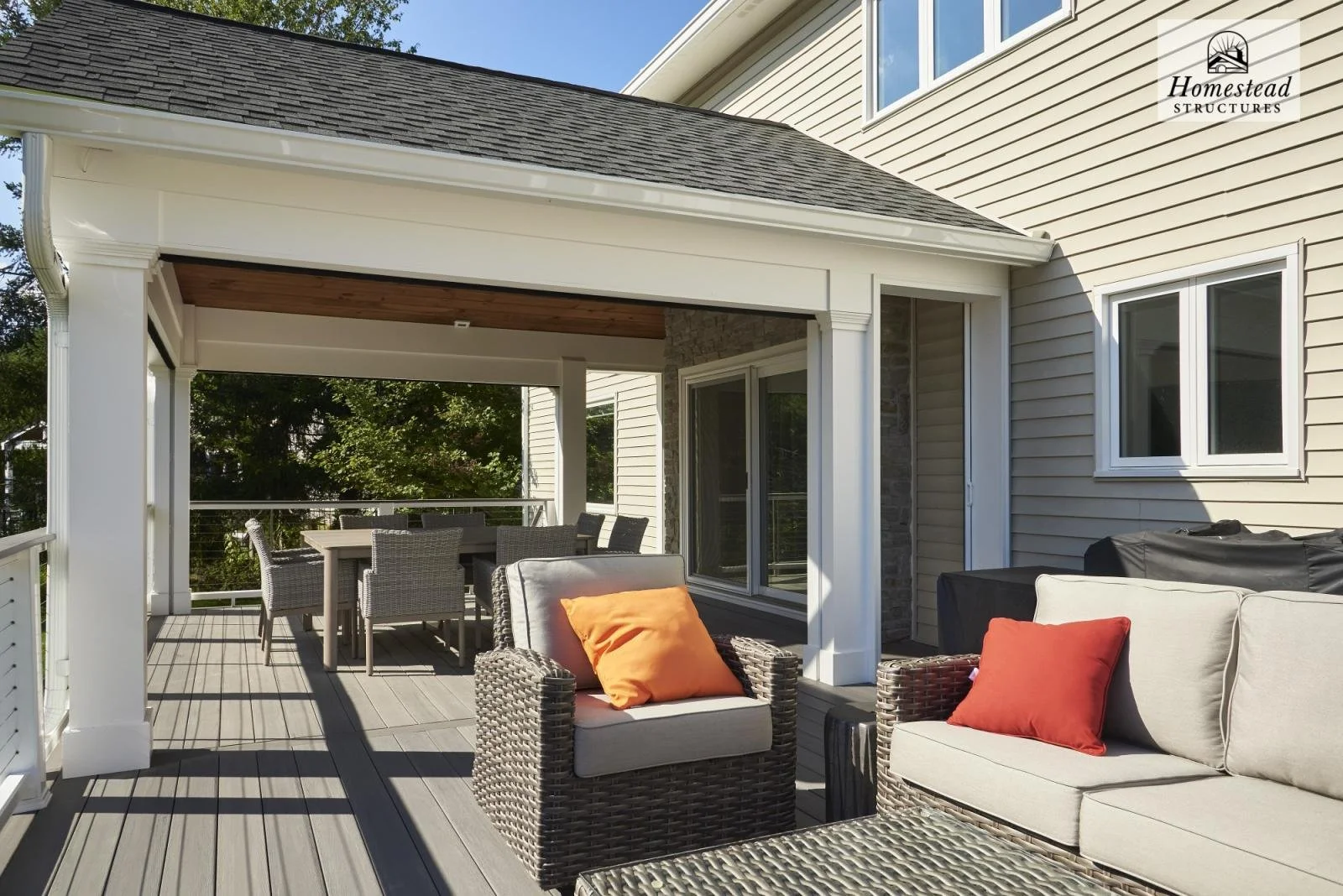 A spacious outdoor deck with wicker furniture and orange cushions, overlooking greenery, attached to a house with beige siding and large windows.