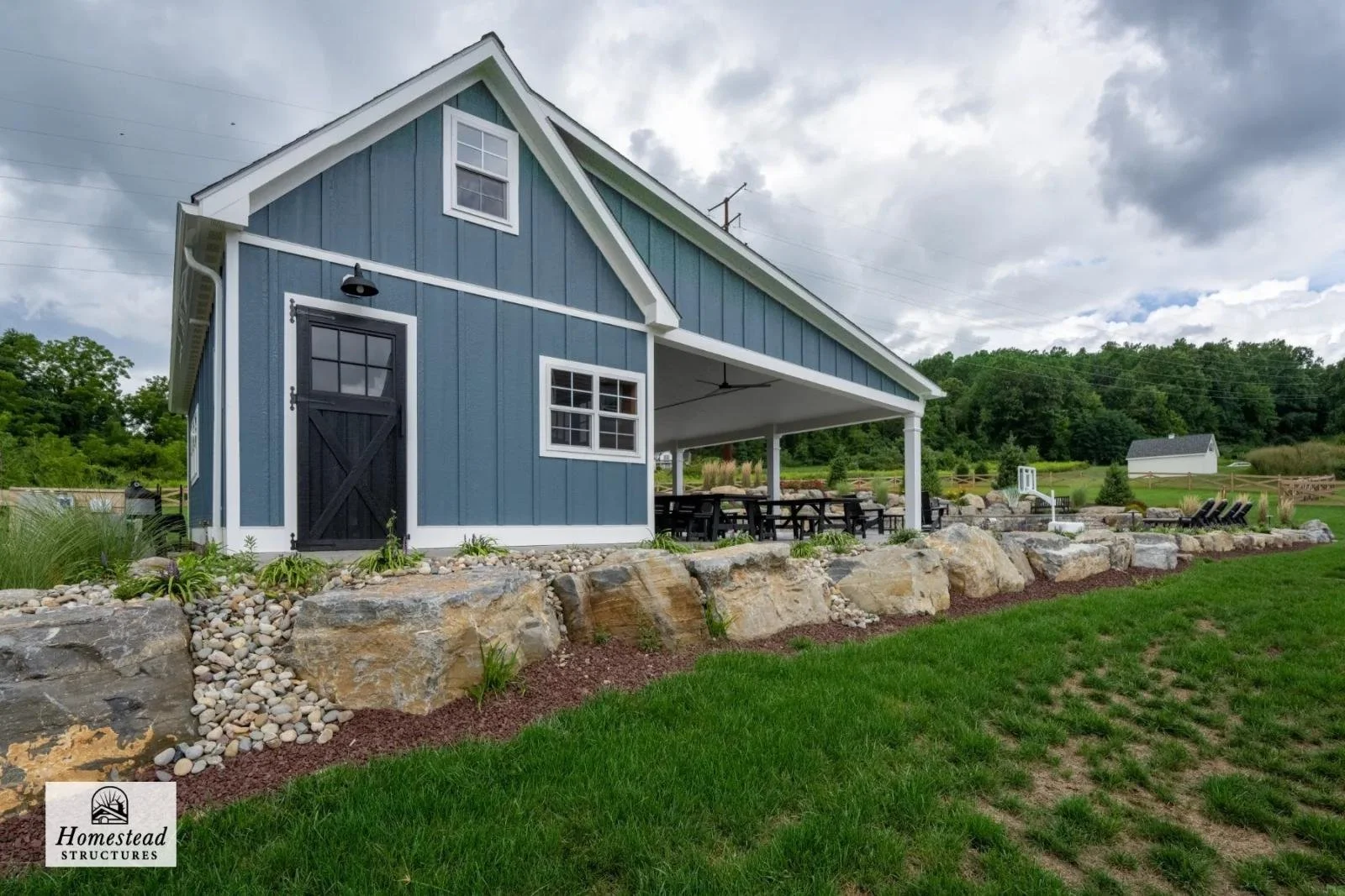Blue barn with black door and white trim, built on a stone foundation, with a covered outdoor dining area, surrounded by a landscaped yard with grass and large rocks, under a cloudy sky.