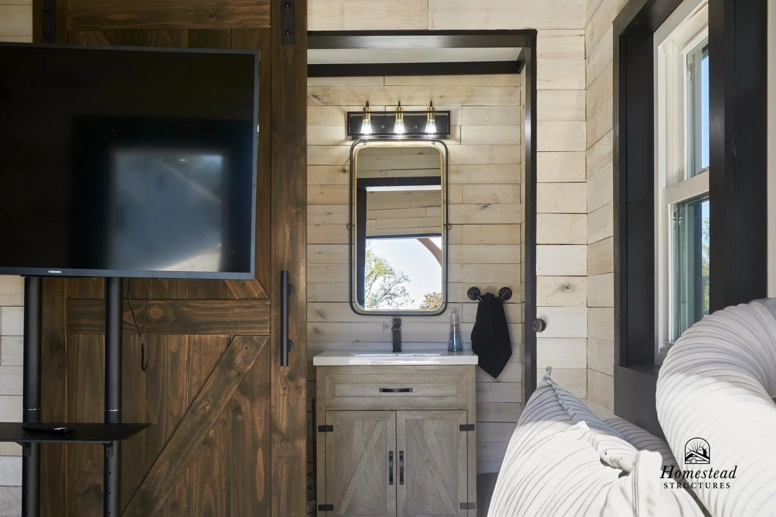 Interior of a rustic-style bathroom with a small vanity, mirror, towel, and window, with a wooden wall and natural light.