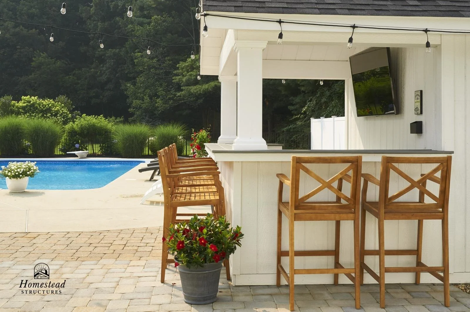 Outdoor pool area with a bar counter, wooden bar stools, potted flowers, and string lights, surrounded by greenery.
