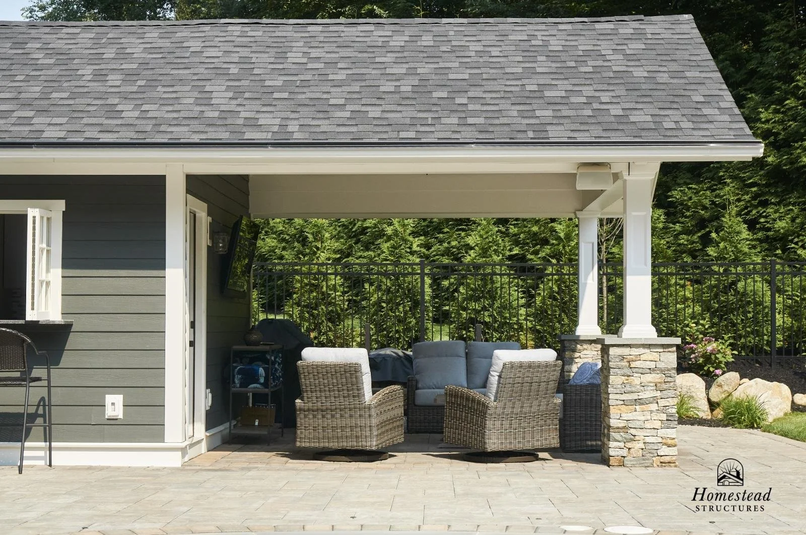 An outdoor patio area with wicker chairs, a cushioned loveseat, and a side table. The patio is covered by a roof and is attached to a house with grey siding. There is a stone pillar supporting the roof and a black fence surrounding the yard with gree