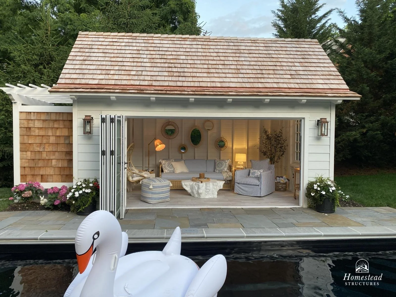 Exterior view of a small, white lakeside cottage with open doors revealing a cozy, decorated living room inside. The cottage has a tan shingle roof, lantern-style exterior lights, and potted plants with flowers outside. In the foreground, there is an