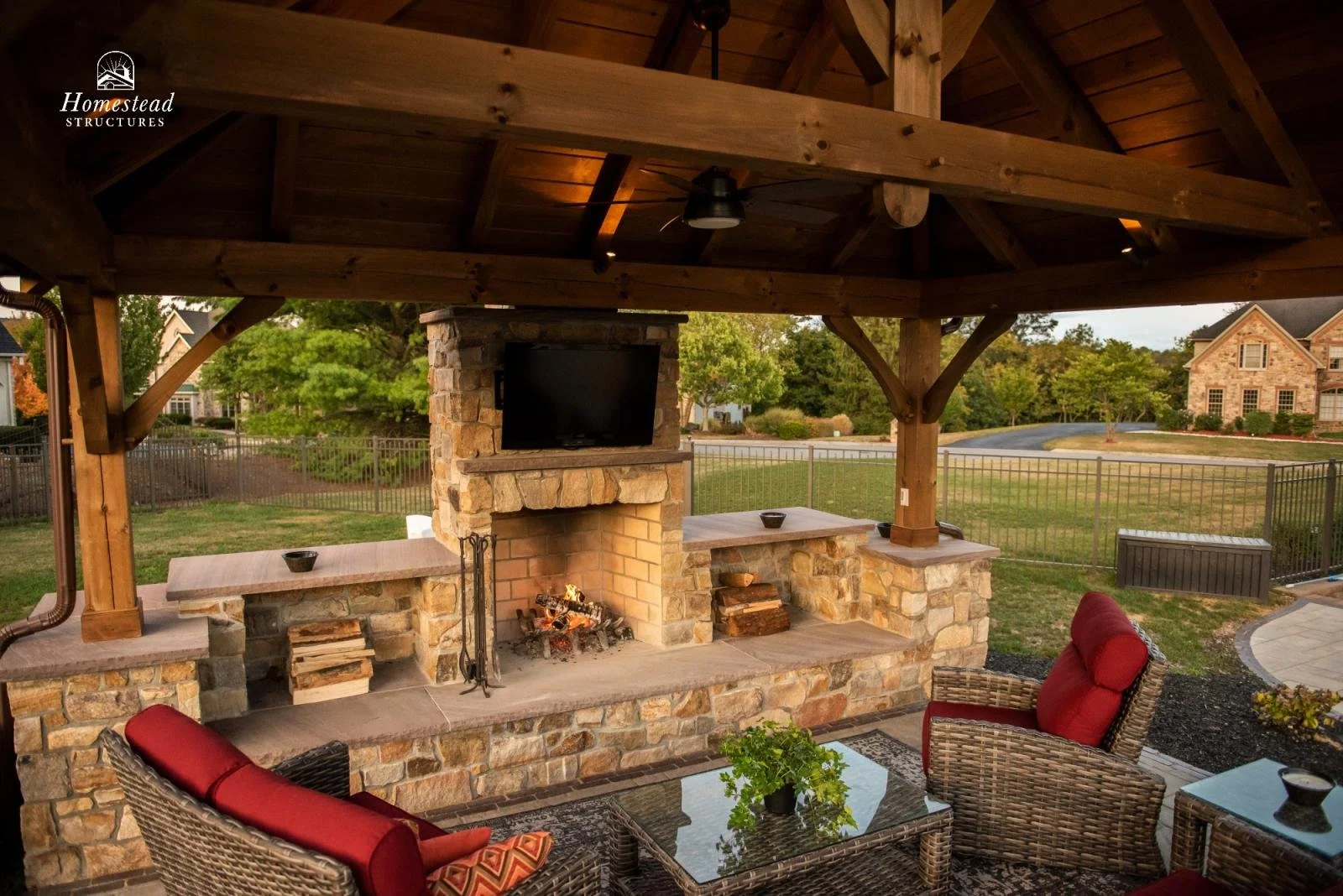 Outdoor living space with a stone fireplace, a mounted television, wicker furniture with red cushions, a glass coffee table with a potted plant, and a wood-finished pavilion roof. Background includes a grassy yard, trees, and neighboring houses.