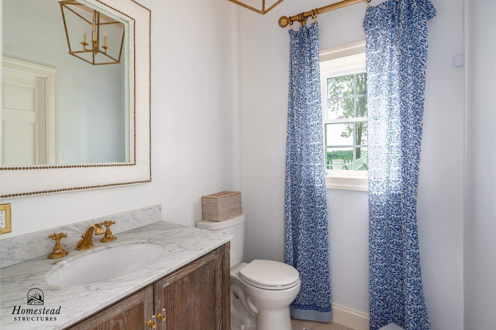 Bathroom with a marble countertop sink and brass fixtures, a mirror with a beaded frame, blue patterned curtains, a window with a view of trees outside, and a wooden cabinet under the sink.