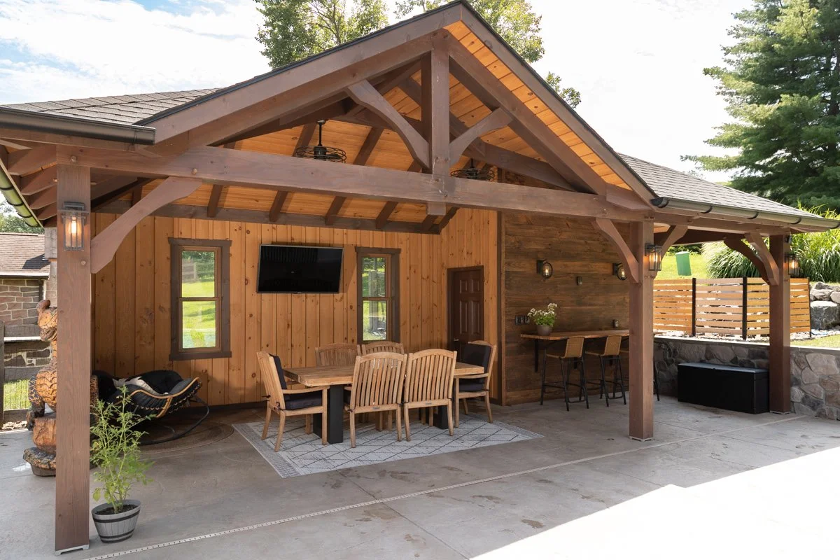 Outdoor patio area with a wooden structure, including a dining table with chairs, a small bar with stools, a hanging TV, and decorative lighting, set against a backdrop of greenery.