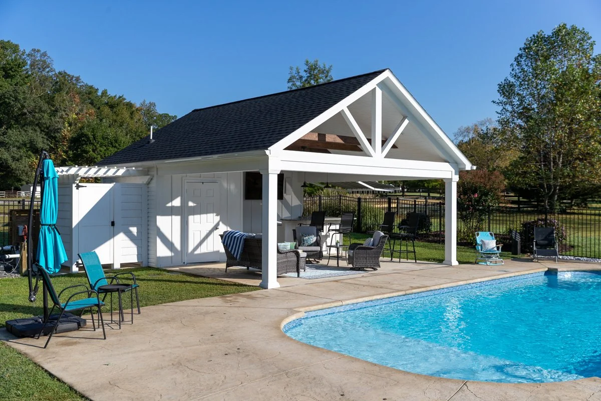 A backyard with a swimming pool, poolside chairs, a covered patio with outdoor furniture, and a small shed or pool house, surrounded by a green lawn and trees.