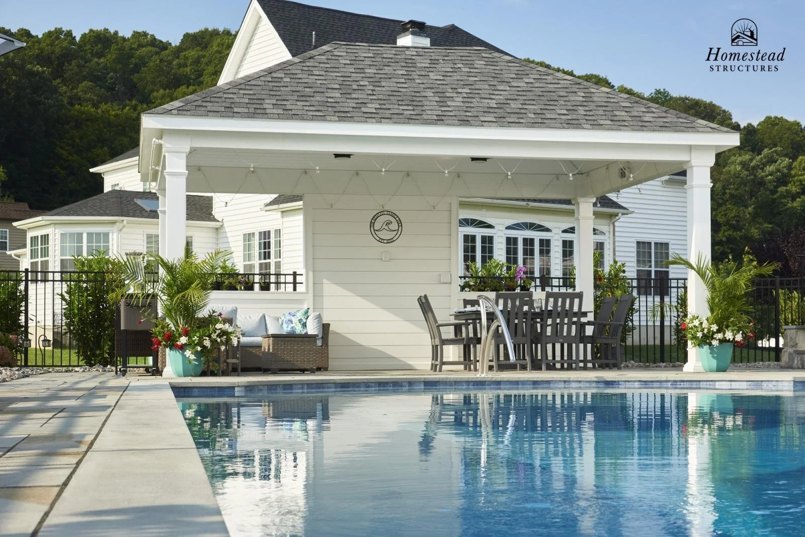 A backyard patio with a swimming pool, outdoor furniture, potted plants, and a white house with large windows in the background.