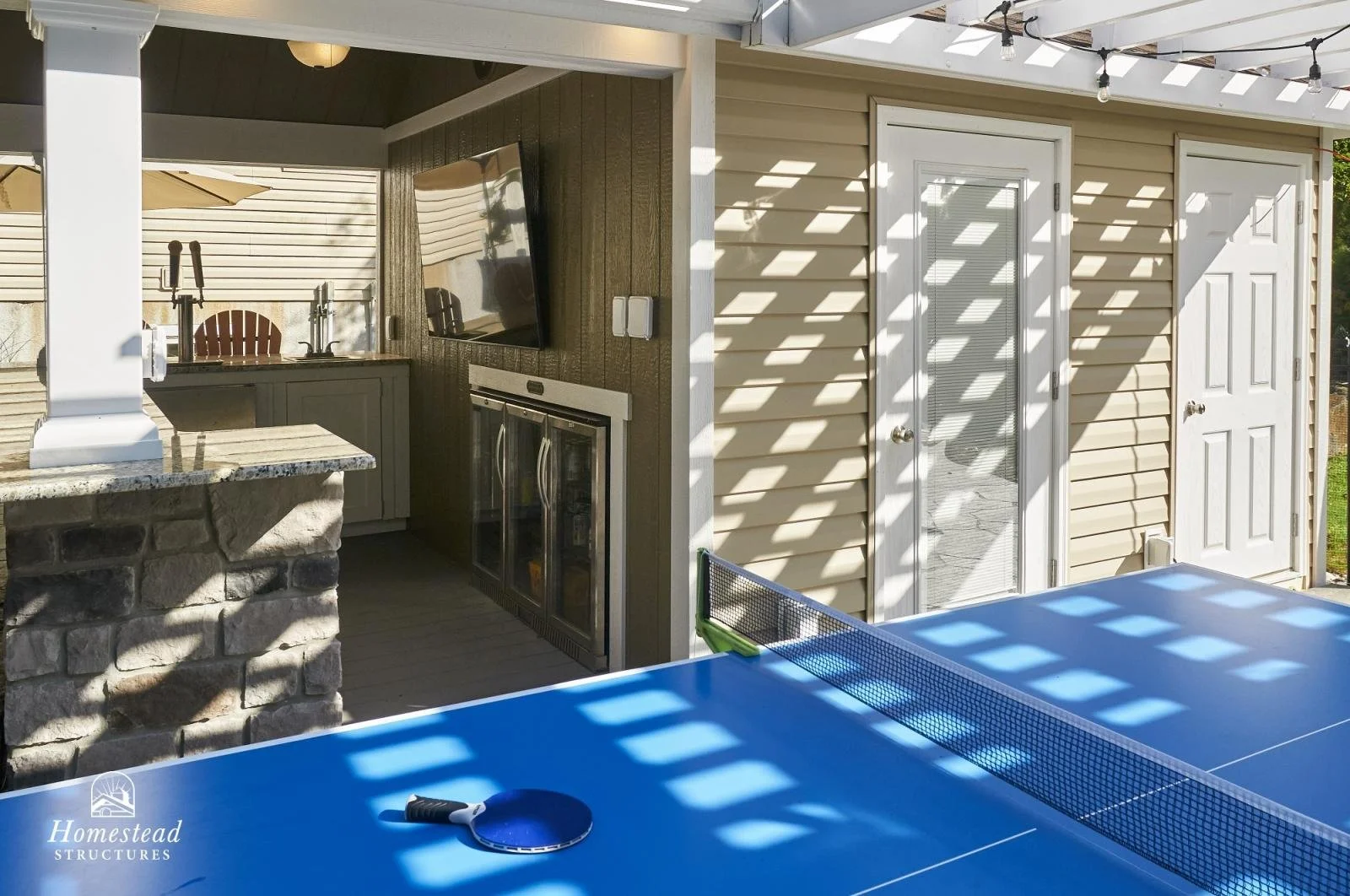 An outdoor patio area with a blue ping pong table and a ping pong paddle. There is a small outdoor kitchen with a sink and wine cooler, a wall-mounted television, and a door leading to an interior space. Sunlight creates a pattern of shadows on the w