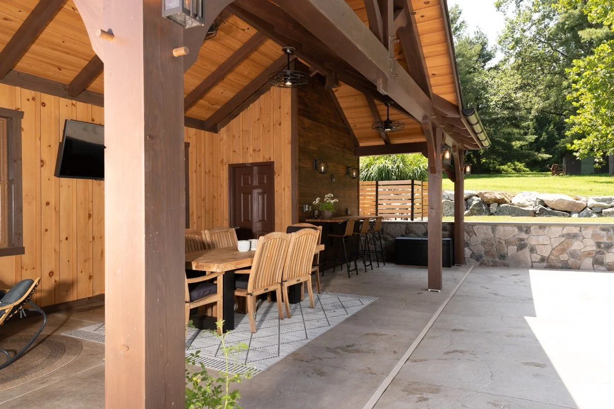Outdoor patio area with wooden walls and ceiling, featuring a dining table with chairs, a bar countertop with bar stools, and a wall-mounted TV, surrounded by greenery and rocks.