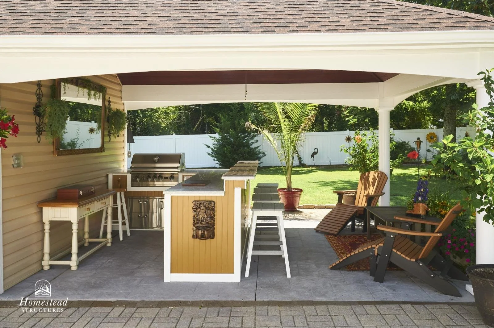 Covered backyard patio with outdoor kitchen, two lounge chairs, a small table, potted plants, and a grassy yard with trees and a white fence in the background.