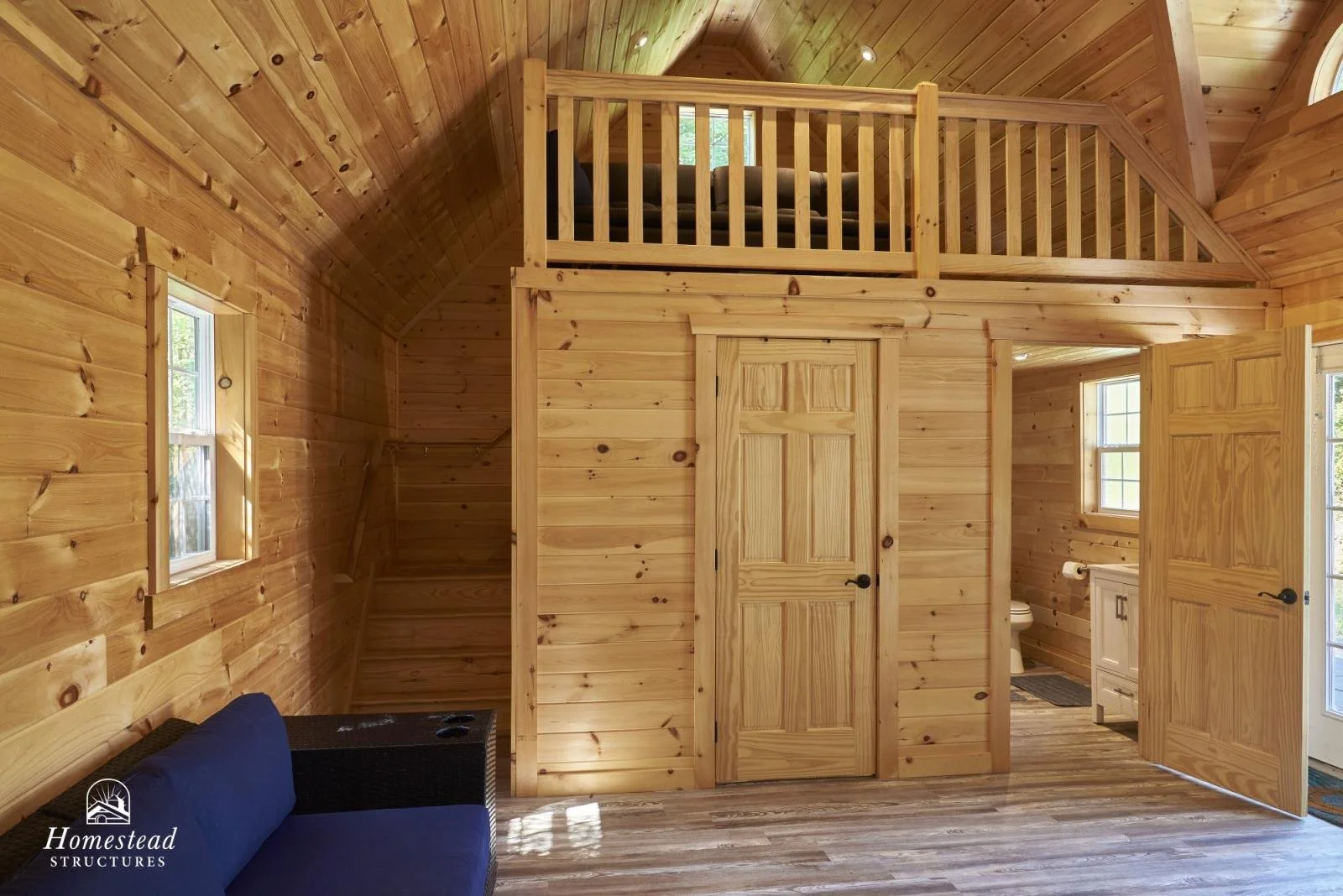 Interior of a wooden cabin with a loft, small windows, a blue couch, and a door leading to a bathroom with a toilet and sink.