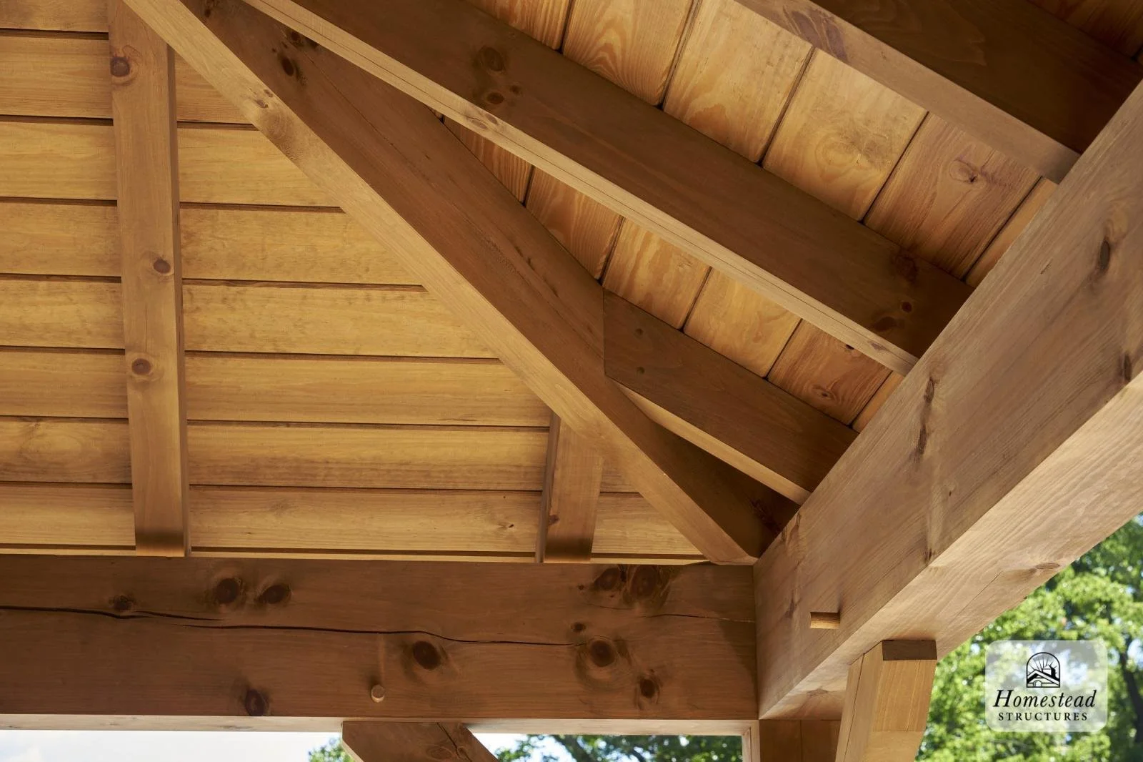 Close-up view of wooden roof structure with beams and plywood, part of a house or building under construction, with sunlight illuminating the wood, and some trees visible in the background.