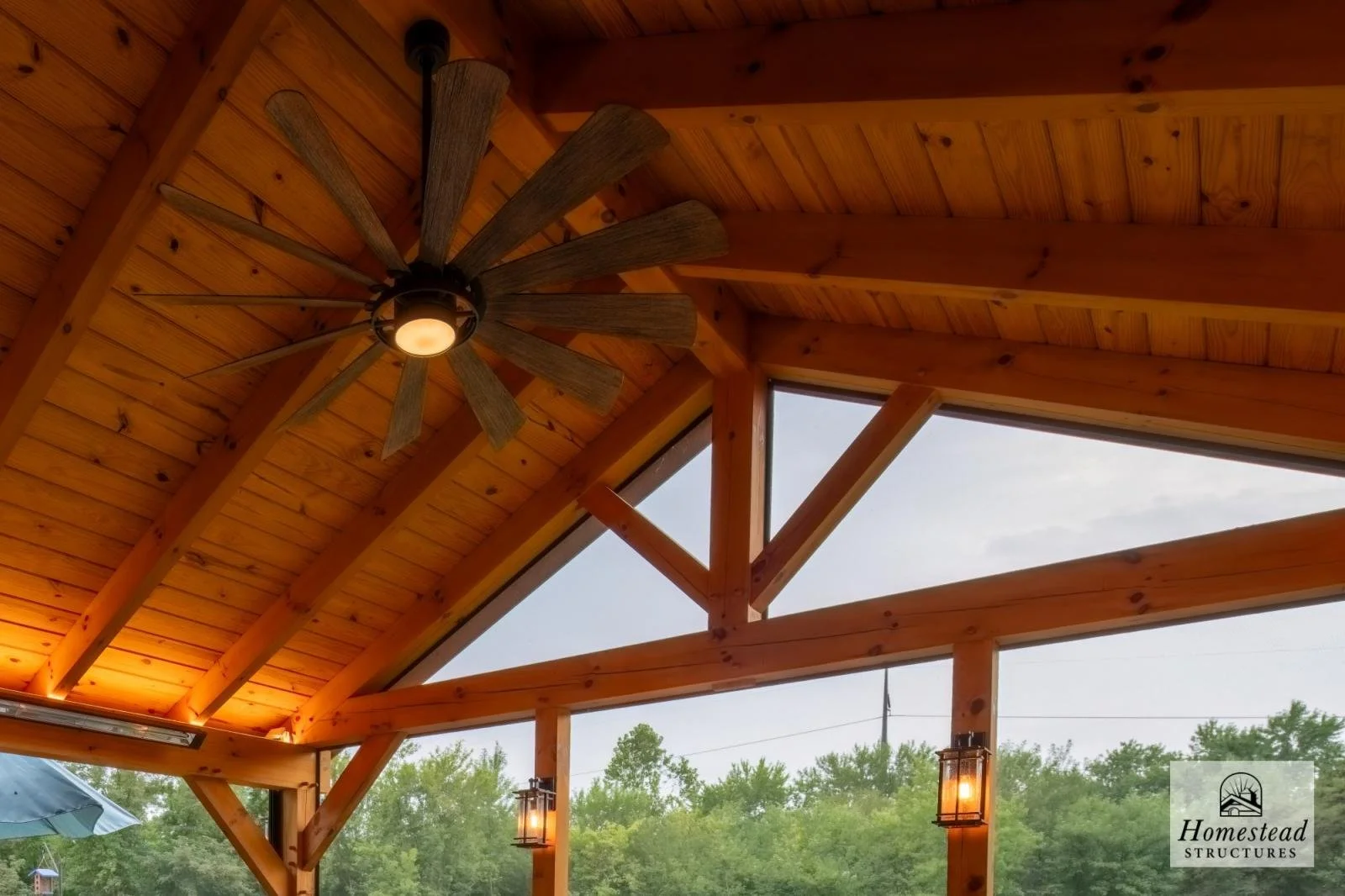 View of a wooden ceiling with a ceiling fan and hanging lantern lights, overlooking a view of green trees outside.