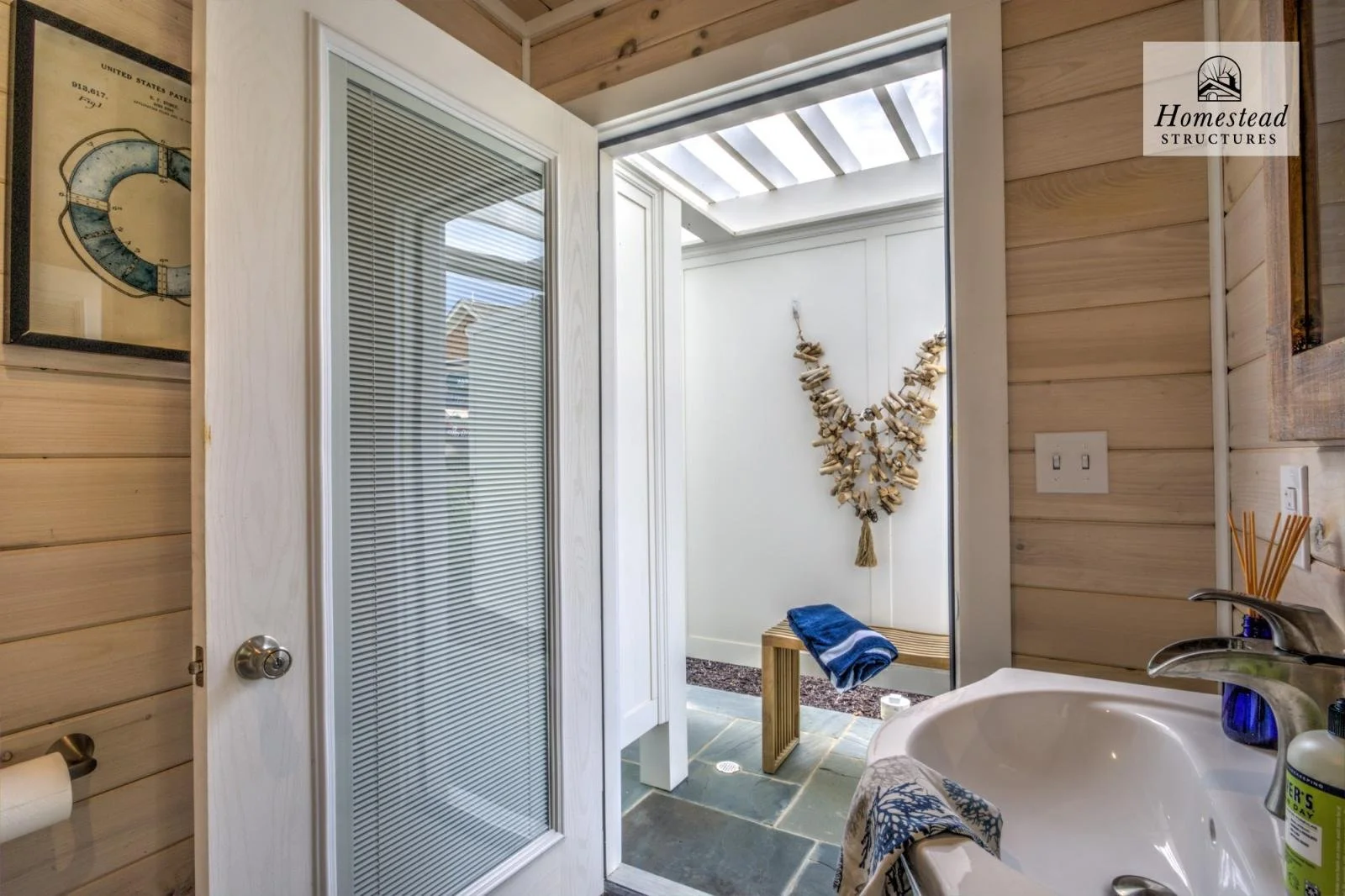 Bathroom with a white door leading to a small outdoor area with a bench, a blue towel, and decorative wall hanging made of seashells. Inside are a sink, toiletries, and wood-paneled walls.