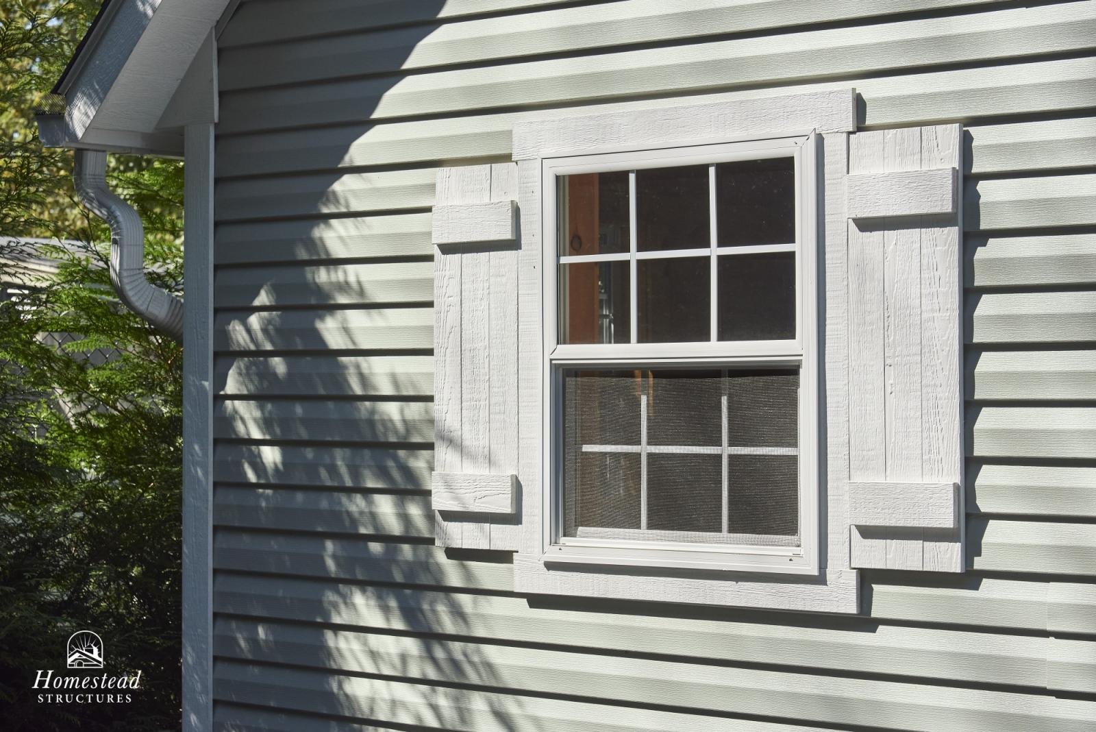 Close-up view of a house siding with a white-framed double-hung window, decorative white shutters, and a metal gutter with a downspout on the side of a house.