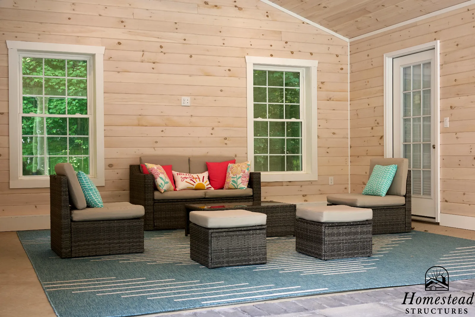 A cozy interior porch with wood-paneled walls, two large windows, and a glass door. There is outdoor furniture including a rattan sofa and chairs with cushions, a coffee table, and a floor rug. The area is well-lit with natural light.
