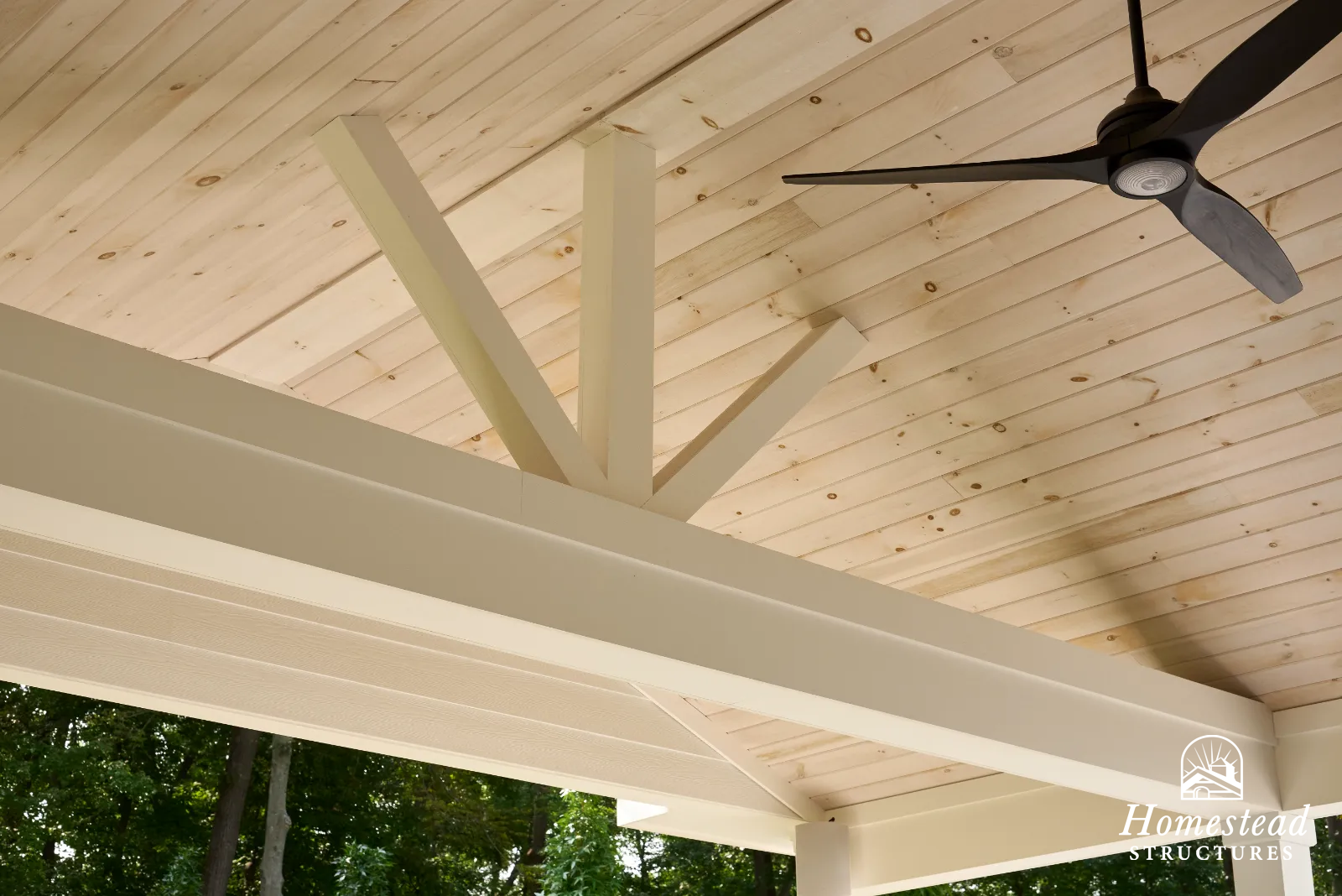 View of a wooden ceiling with a ceiling fan, beams, and greenery outside.