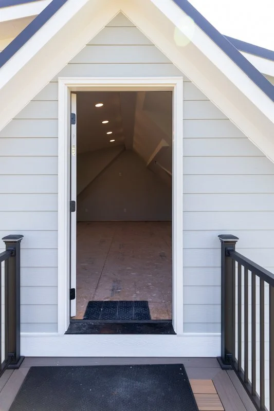 View of a small attic or loft space through an open door from a balcony or deck, with black railings and a doormat outside.