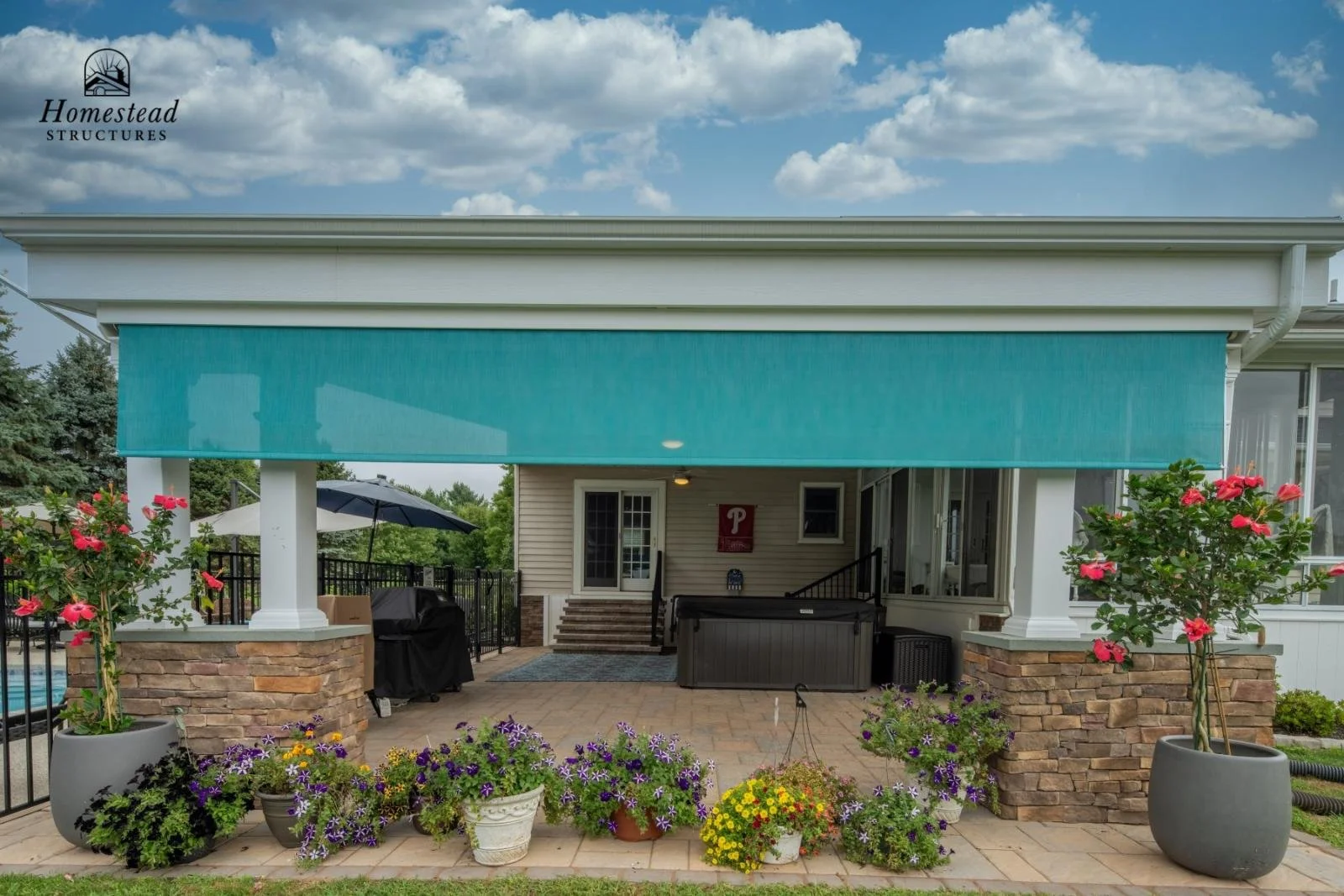 Backyard patio with potted pink hibiscus and purple petunias, stone pillars, hot tub, patio umbrella, and a house with a covered porch and a blue retractable awning.
