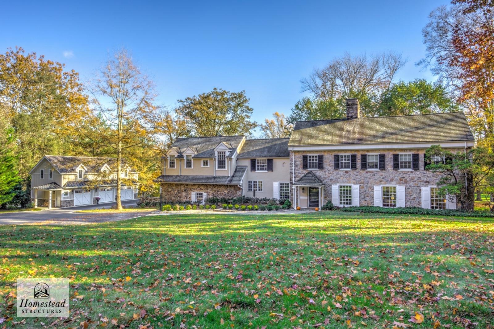 Large house with a stone and beige exterior, surrounded by trees with fall foliage, a front lawn with fallen leaves, and a blue sky.