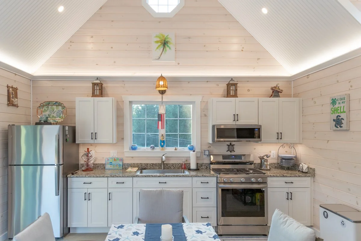 Kitchen with white cabinets, granite countertops, stainless steel refrigerator and stove, window above sink, light wood walls, and tropical decor accents.