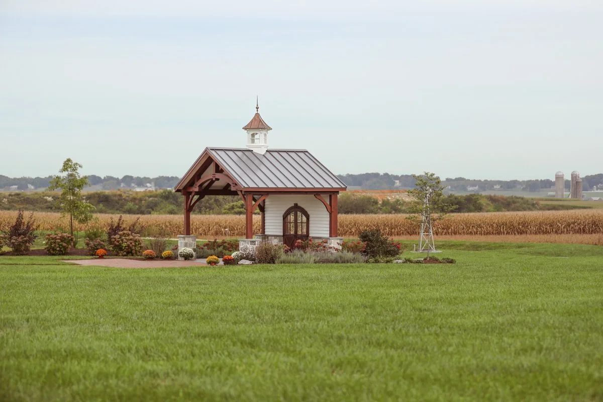 A small decorative building with a brown roof and white walls, situated in a lush green field with flower beds and trees, with a rural landscape and silos in the background.