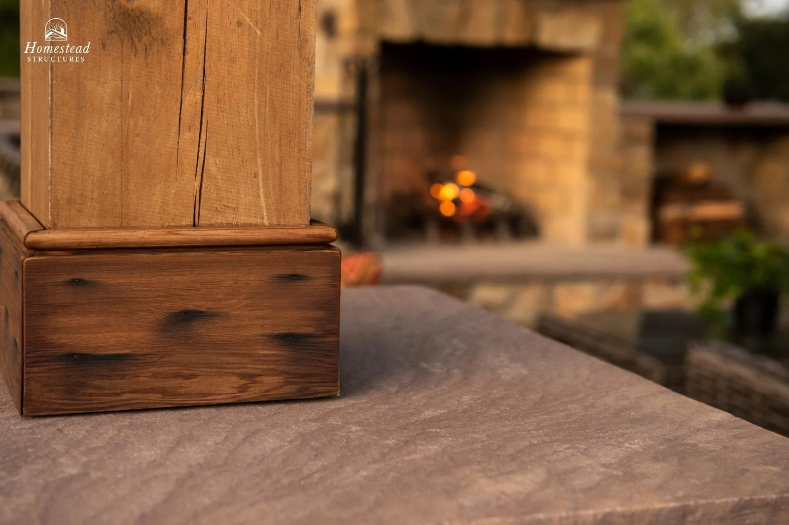 Close-up of a wooden post and a wooden square block on a table in front of an outdoor brick fireplace with a fire burning, with blurred trees and outdoor furniture in the background.