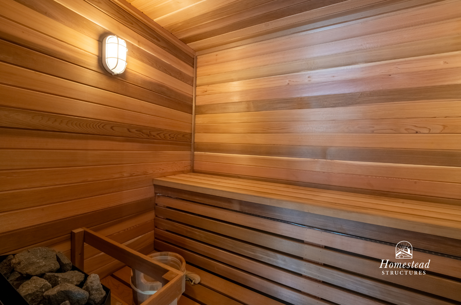 Inside a wooden infrared sauna with wood paneling, a light fixture on the wall, a bench, and sauna stones with a bucket and scoop in the corner.