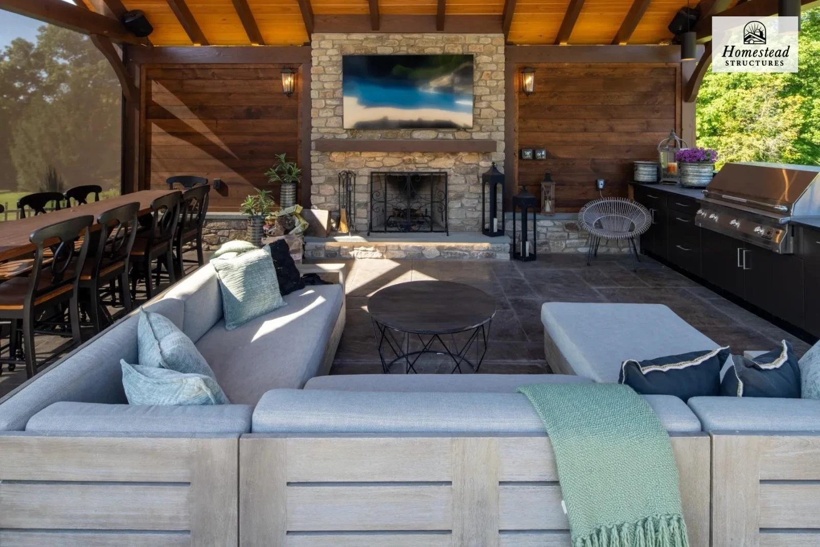 Outdoor living space featuring a stone fireplace with a mounted flat-screen TV, surrounded by couches, a coffee table, and string lights, with a dining table to the left and a barbecue grill on the right.