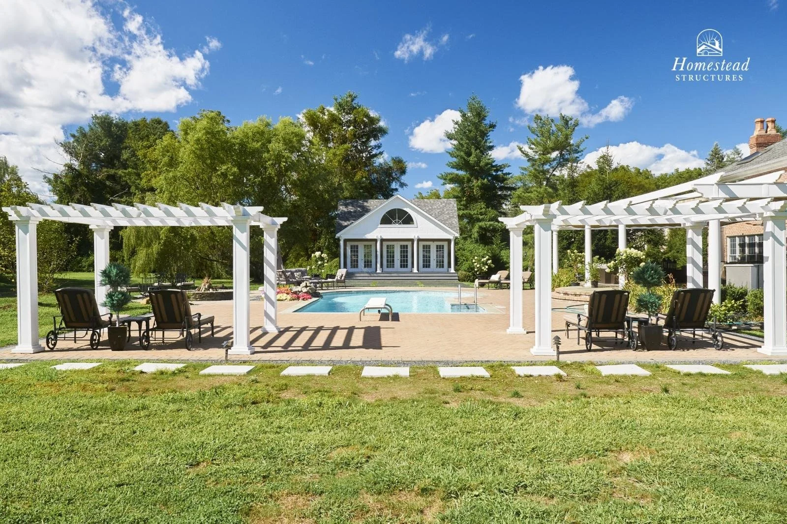 A backyard with a swimming pool, surrounded by white pergolas with lounge chairs. In the background, there is a small white building with large windows and a deck. Tall trees and a blue sky with scattered clouds are visible.