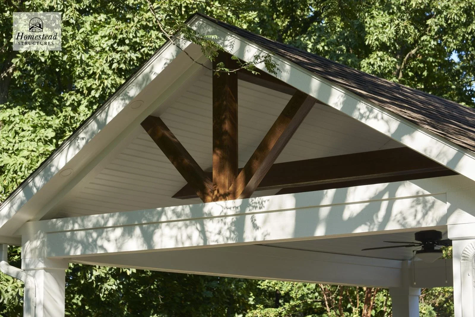 Close-up of a white house porch roof with dark brown support beams, surrounded by green trees, casting shadows. Homestead Structures logo in the top left corner.