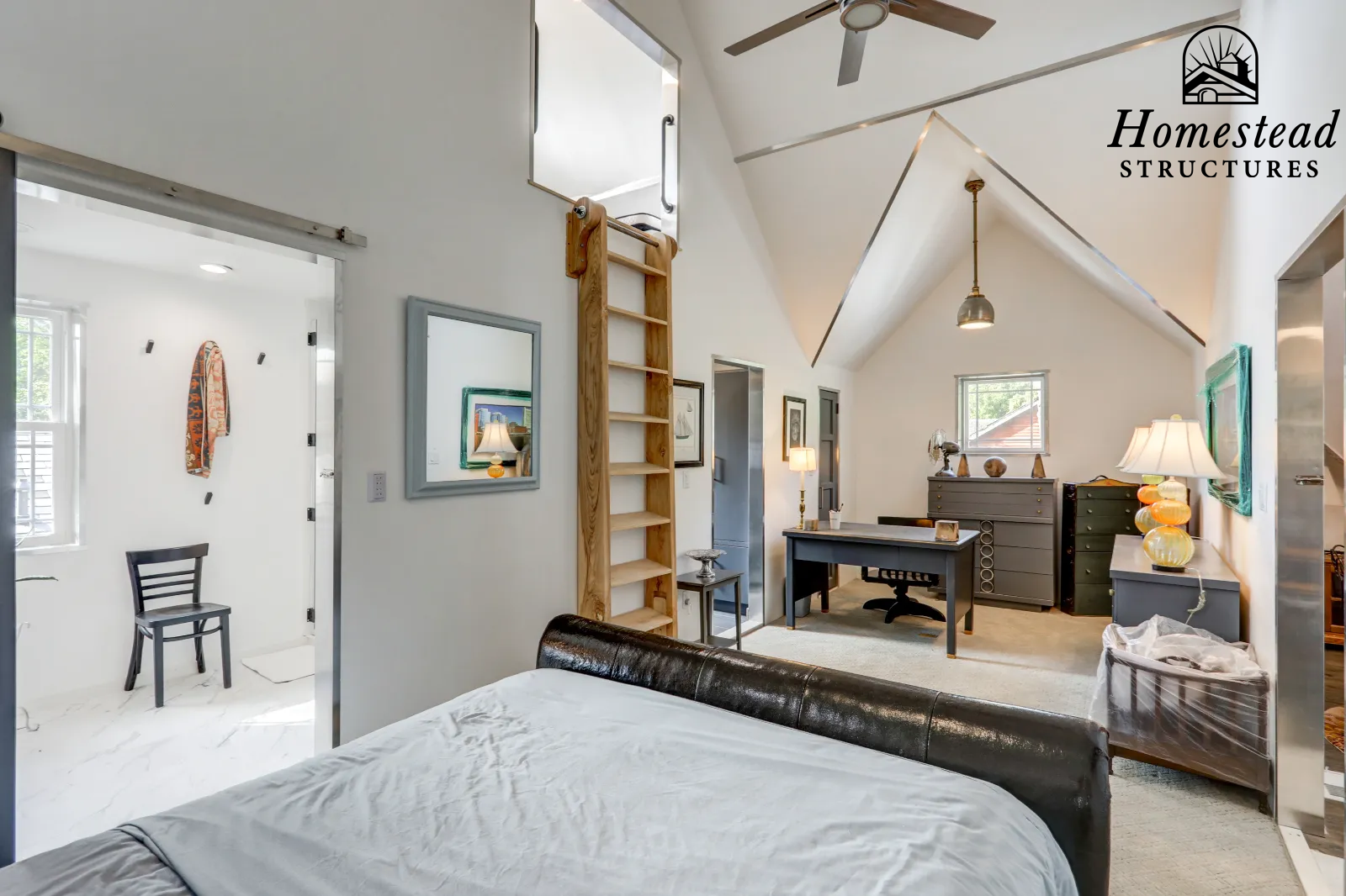 Interior view of a bedroom with a high ceiling, a bed with a leather headboard, a wooden ladder leading to an attic space, a desk with a lamp, and various decorative items and lamps. There is an adjacent bathroom visible through an open door.