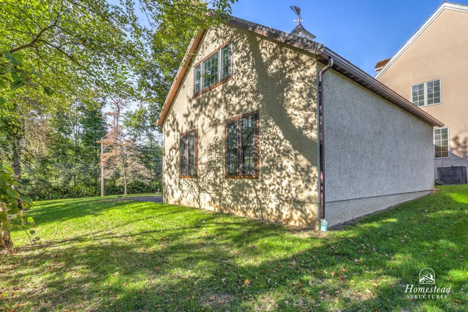 A two-story house with a textured beige exterior and brown window trim, surrounded by green grass and trees, with shadows cast on the house by the leaves.
