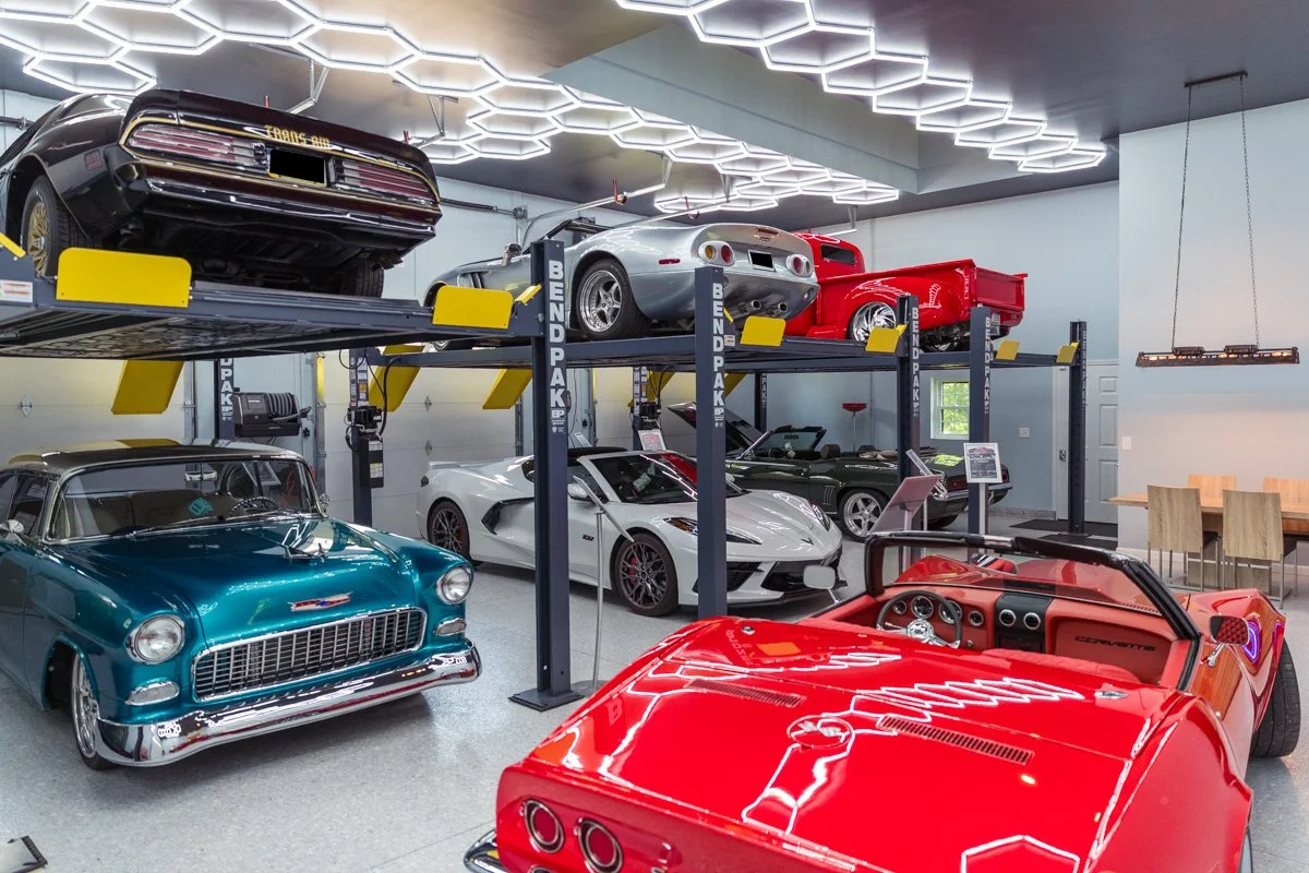 A collection of classic and exotic cars in a showroom or garage, including a blue vintage car, a white sports car, a black car on an upper lift, a silver sports car, and a red pickup truck, with modern hexagonal ceiling lighting.