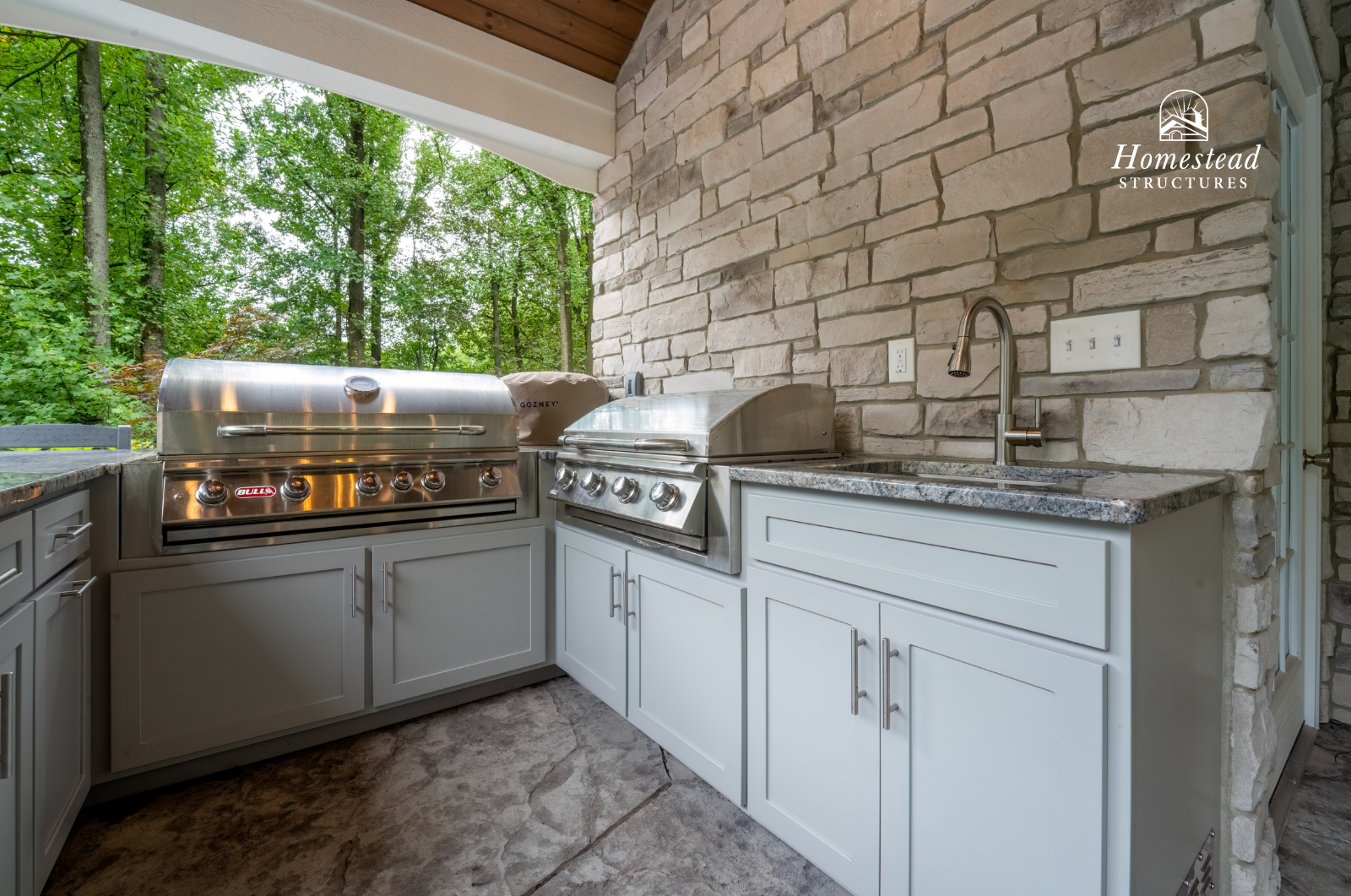 Outdoor kitchen with gray cabinets, granite countertops, and two stainless steel grills, set against a stone wall with trees in the background.