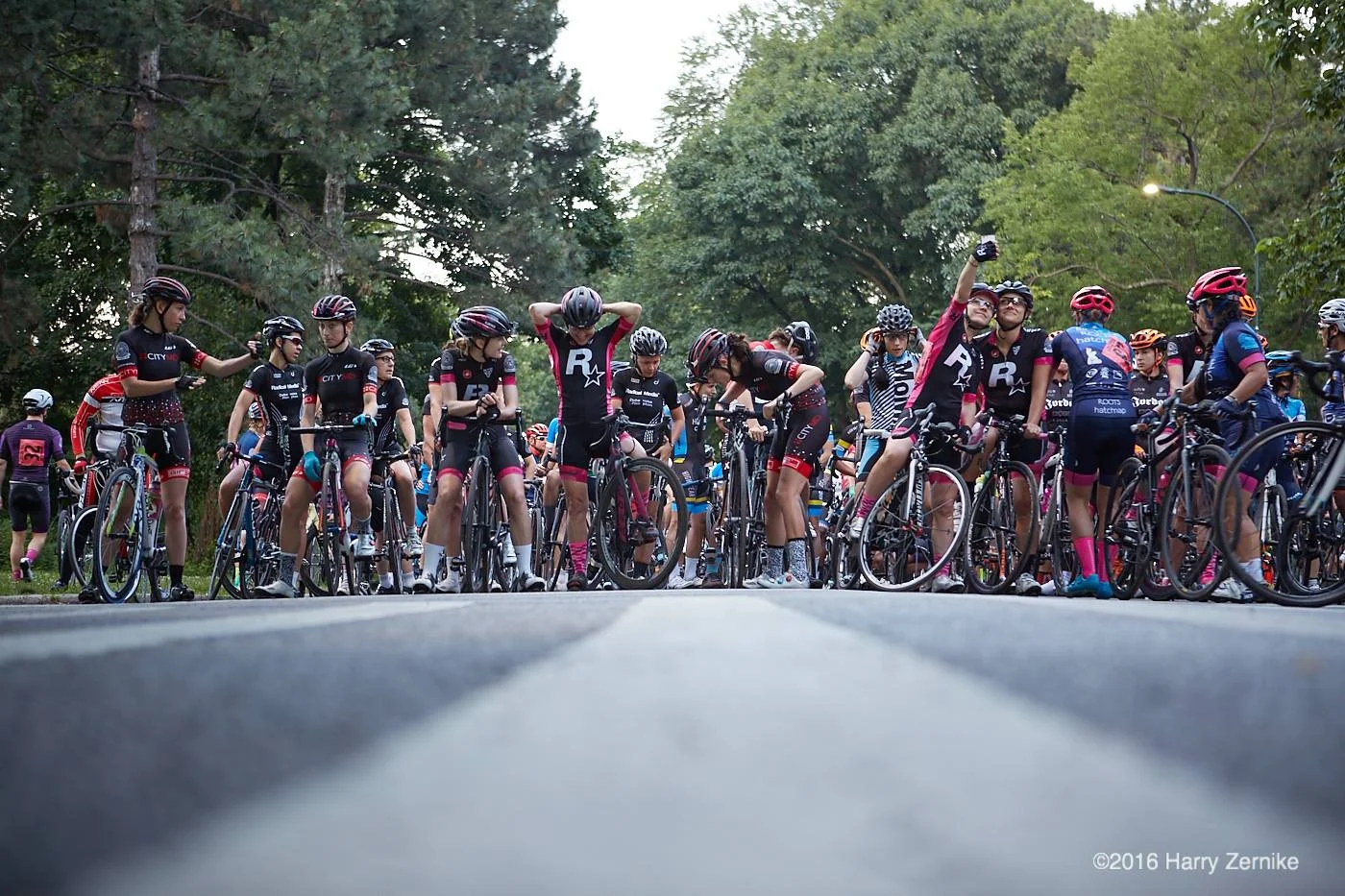 Group of cyclists gathered on a road, preparing for a race, with trees in the background.