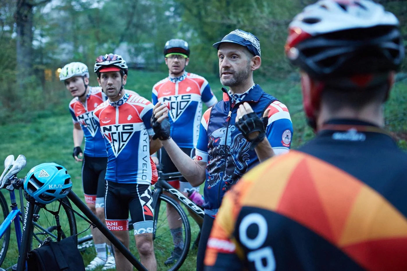 Group of cyclists in matching jerseys standing outdoors, one speaking to the others, with bicycles around them.