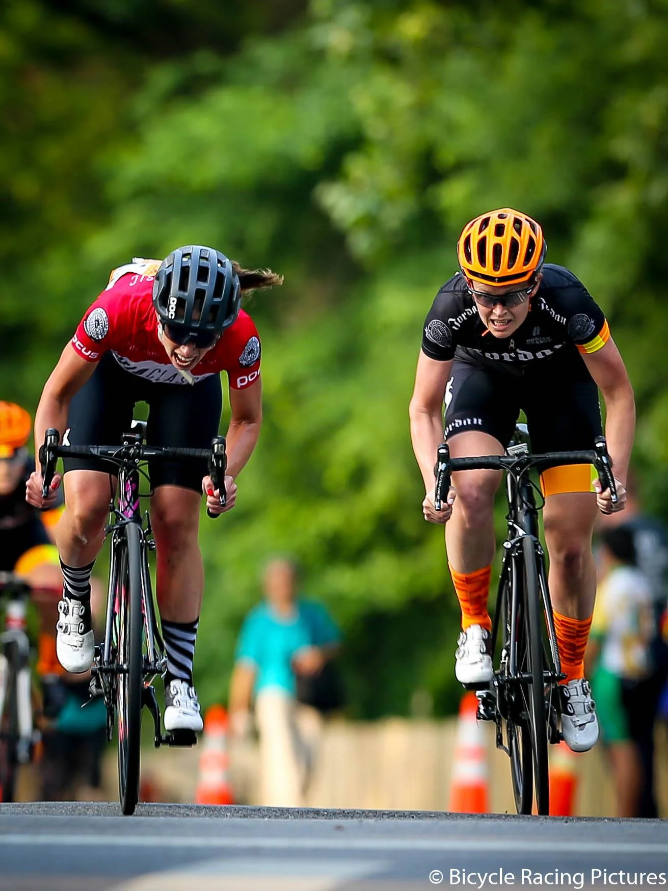 Two cyclists racing intensely on a road, wearing helmets and cycling gear, with blurred green background.