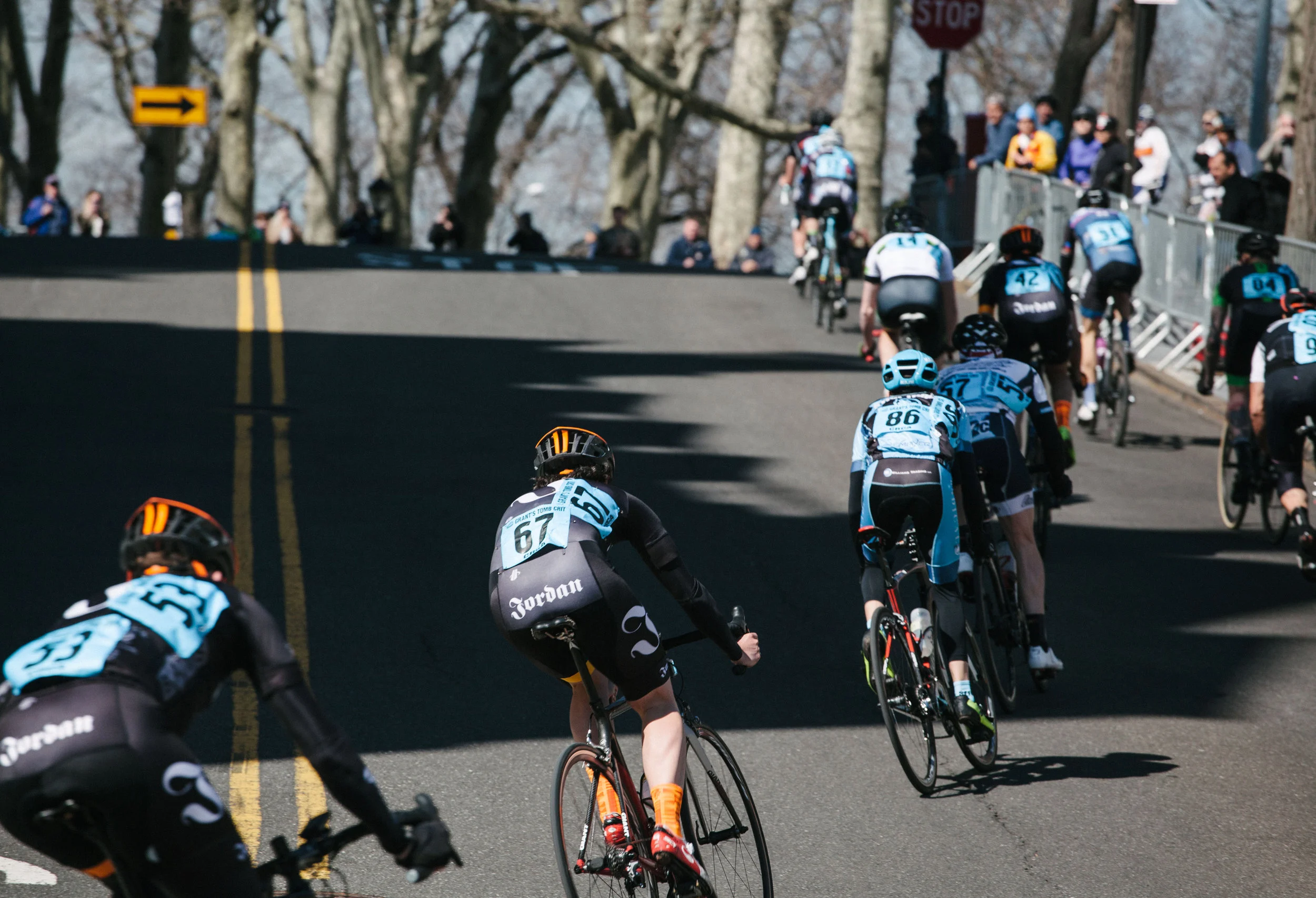 Cyclists in a road race riding uphill with spectators on the side, numbered jerseys visible, and trees in the background.