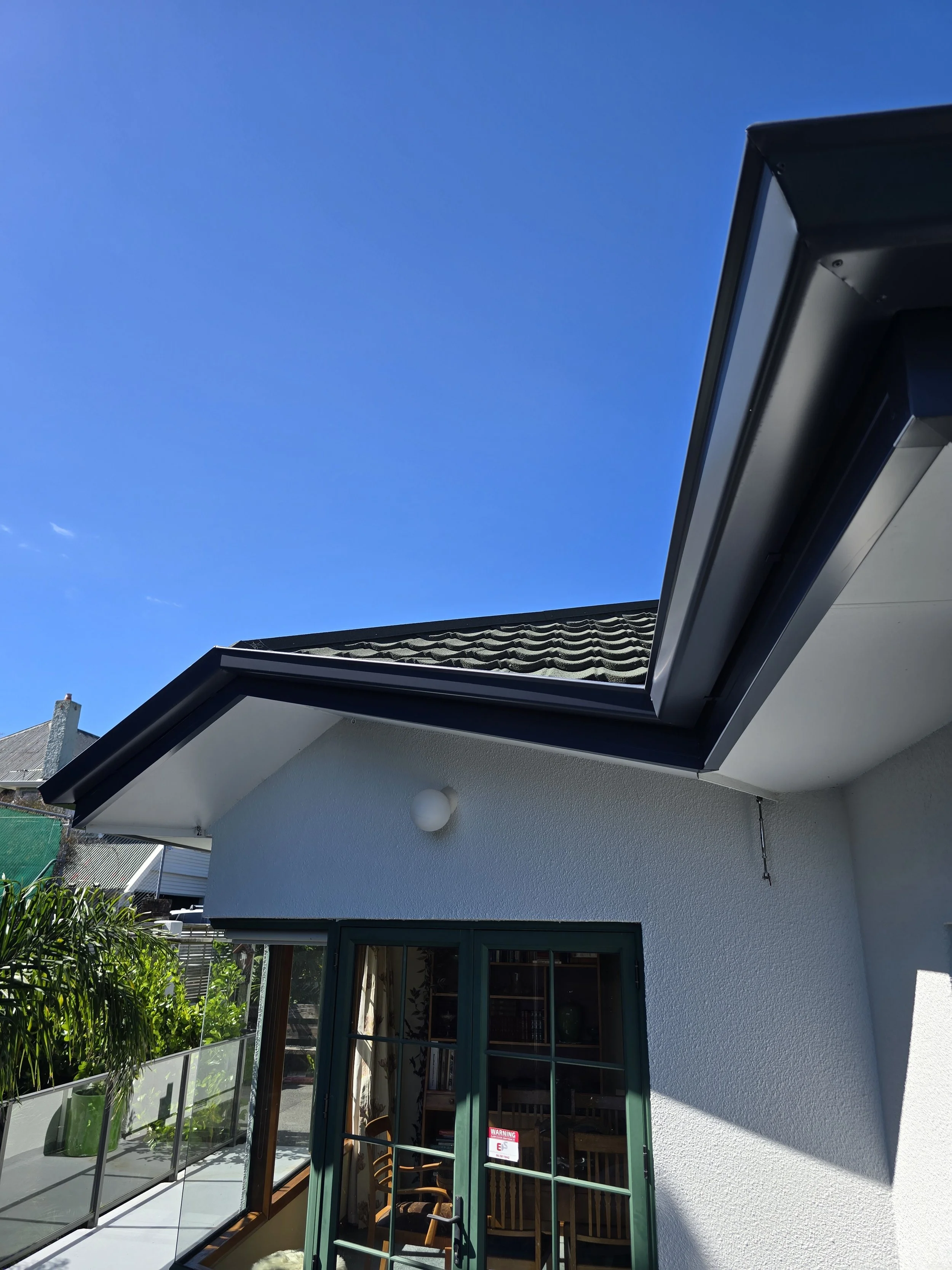 A house with a glass door, white exterior wall, and an open skylight on the roof. There is a sign on the door and outdoor furniture inside.