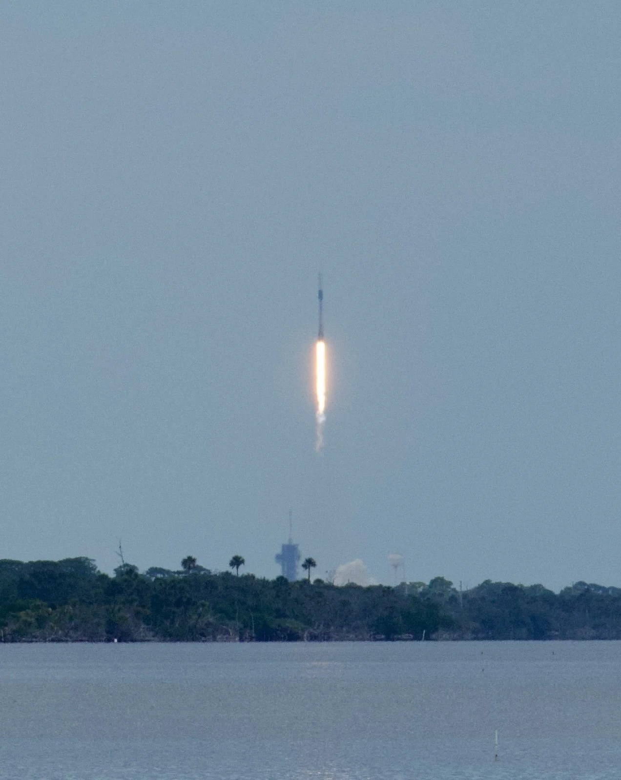 Rocket launching into the sky over a body of water with trees and a building in the background.