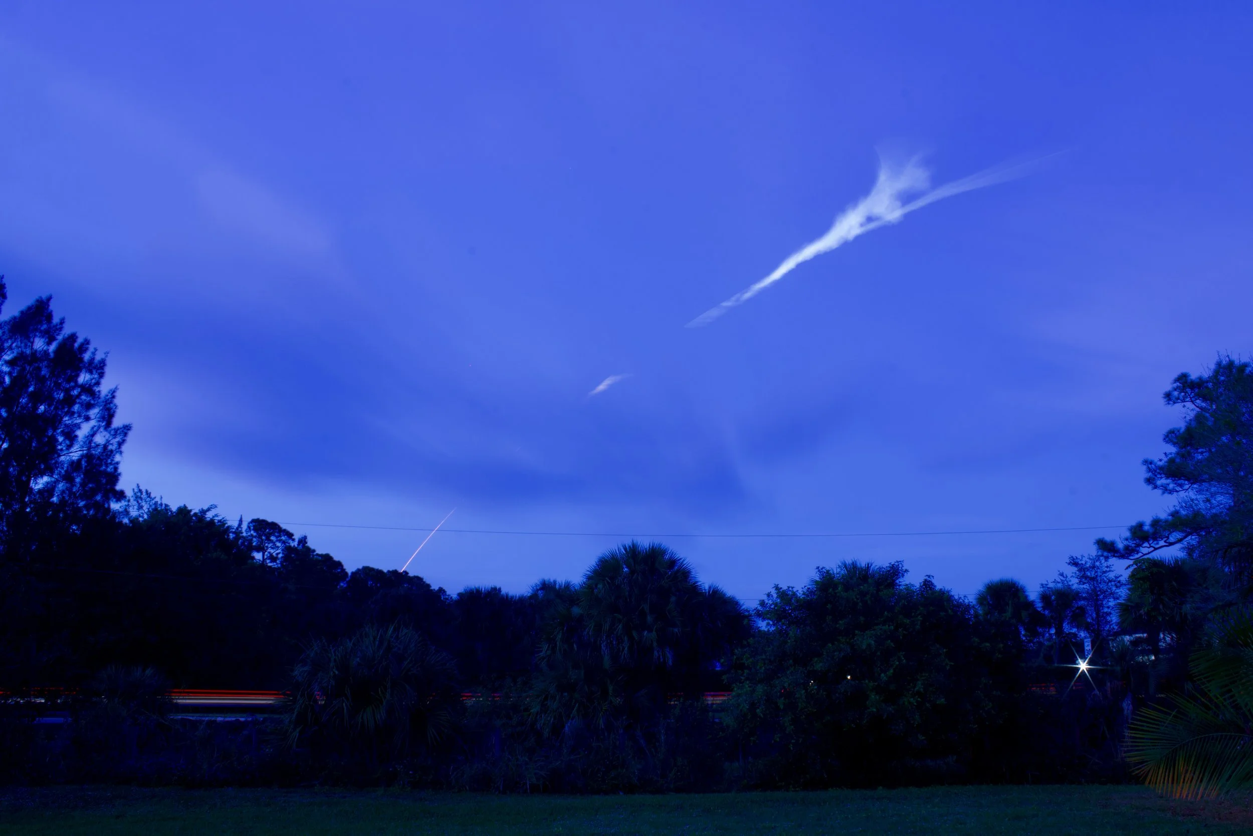 Nighttime scene with dark trees and bushes under a deep blue sky with streaks of white clouds and distant light trails.