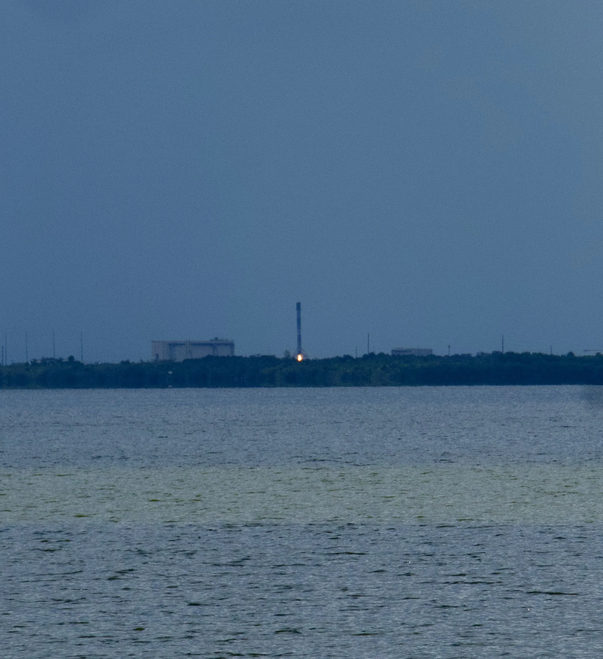 View of a body of water with a distant landscape, including a tall lighthouse and industrial buildings on the horizon at dusk or dawn.