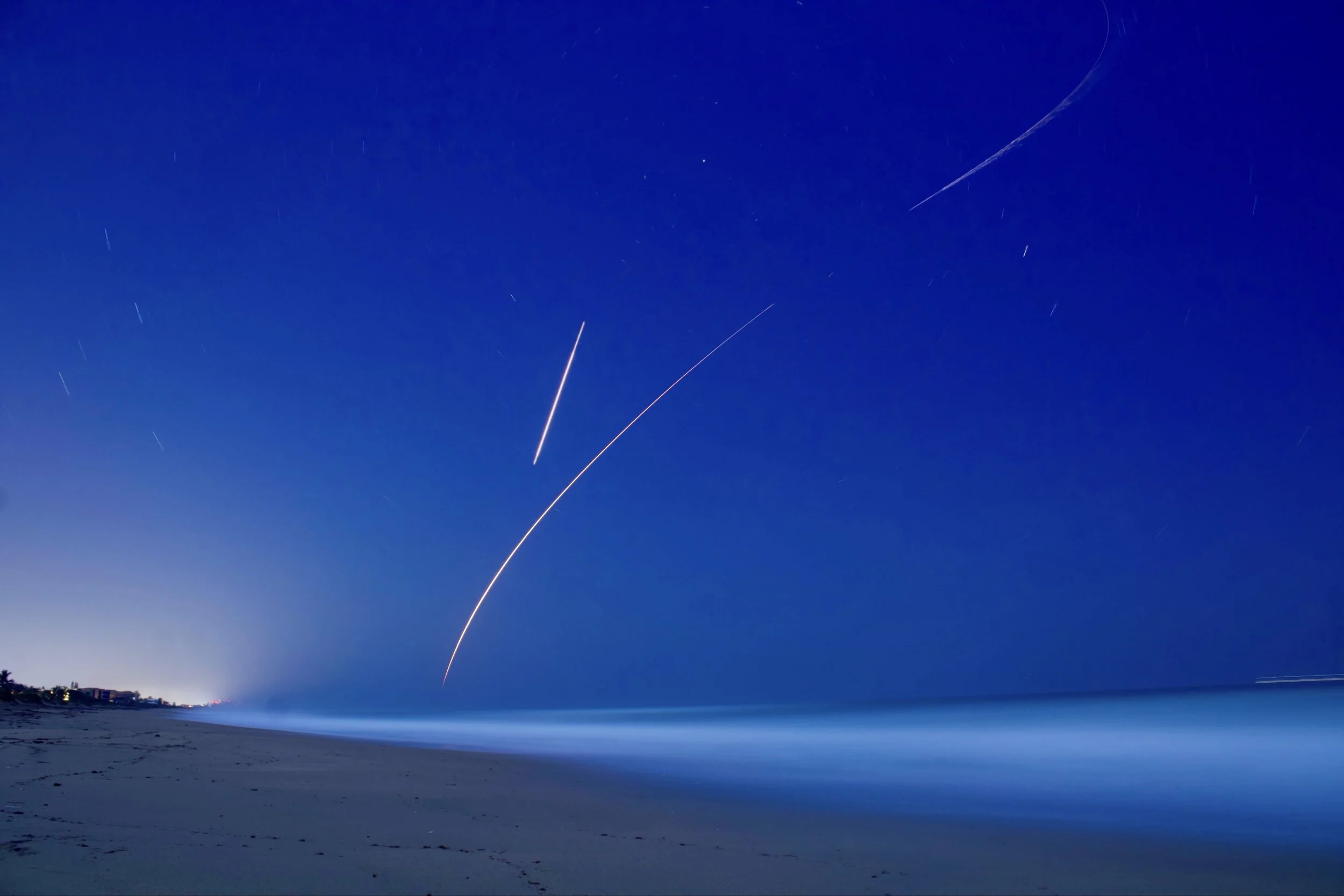 Nighttime sky over the ocean with star trails and streaks from a shooting star, with a sandy beach in the foreground.