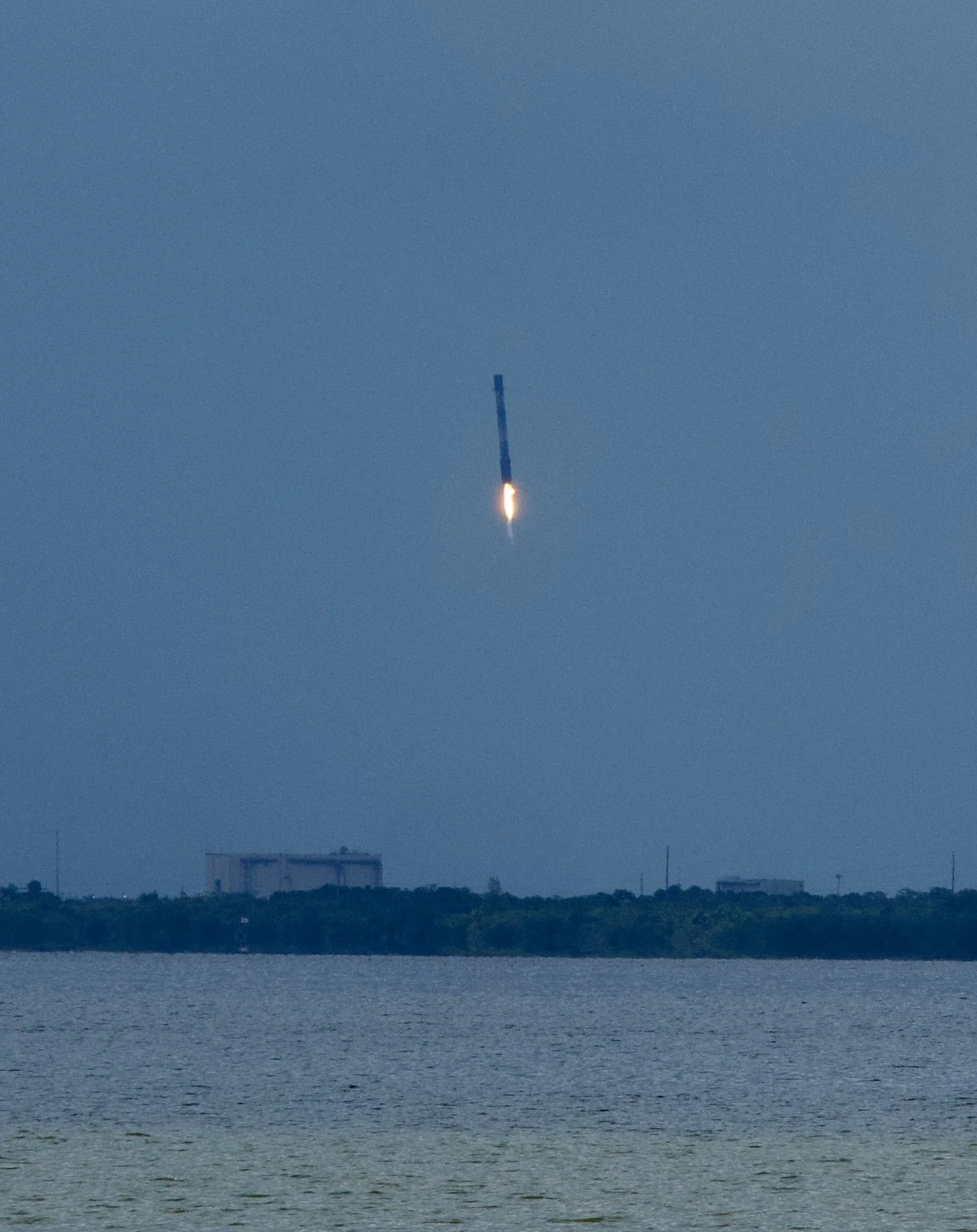 A rocket launching into the sky over a body of water with industrial buildings in the background.