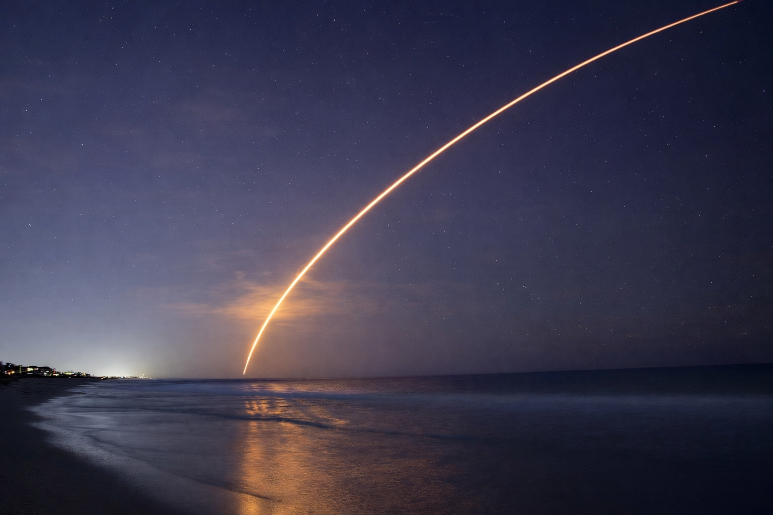 Nighttime scene of a rocket launch over the ocean, with a trail of bright light streaking across the sky and reflections on the water.
