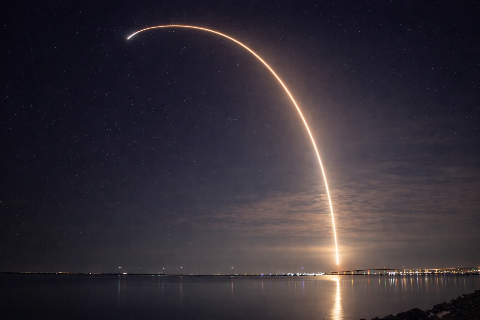 Night sky with stars and a bright streak of light from a rocket launch over a body of water with city lights in the distance.