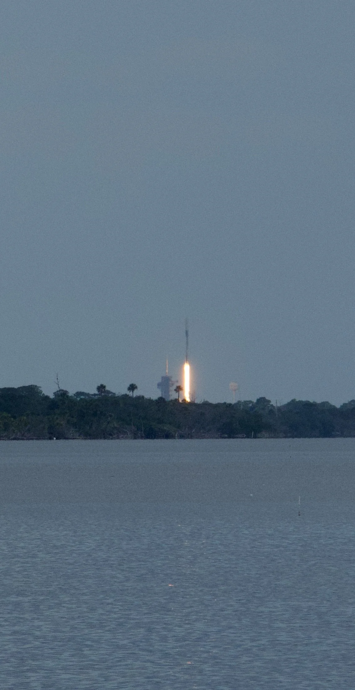 A rocket launching from a shoreline with a water body in the foreground and a small, wooded island in the background.